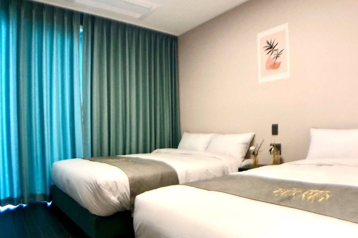 Habitación de hotel con dos camas, un ventanal con cortinas azules y una lámpara fijada a la pared.