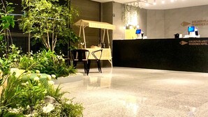 Zona de recepción con mostrador, área de descanso con sillas y un pequeño jardín con plantas y un árbol.