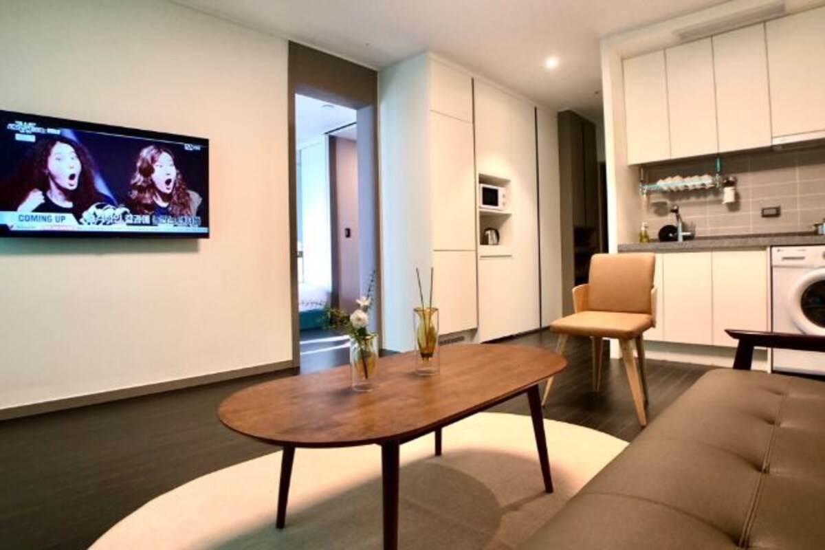 Una sala de estar moderna con un televisor de pantalla plana, un sofá, una mesa de centro y una cocina con armarios y electrodomésticos.