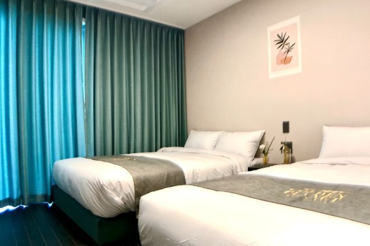 Una habitación de hotel con dos camas, un ventanal con cortinas, una obra de arte en la pared y una mesita de noche con un florero.