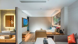 A modern hotel room with a bathroom, a seating area with a coffee table, and a large painting on the wall.