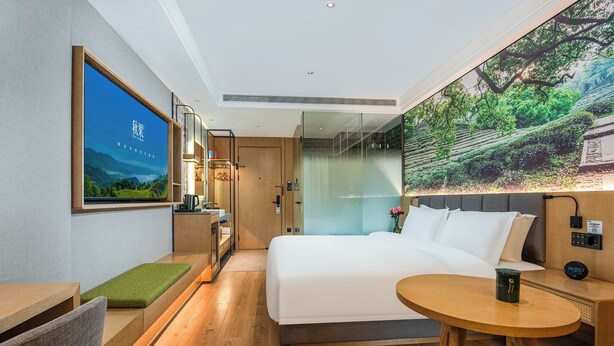 A modern hotel room with a large bed, a small round table, a desk with a chair, a shower area, and a large window with a scenic view.