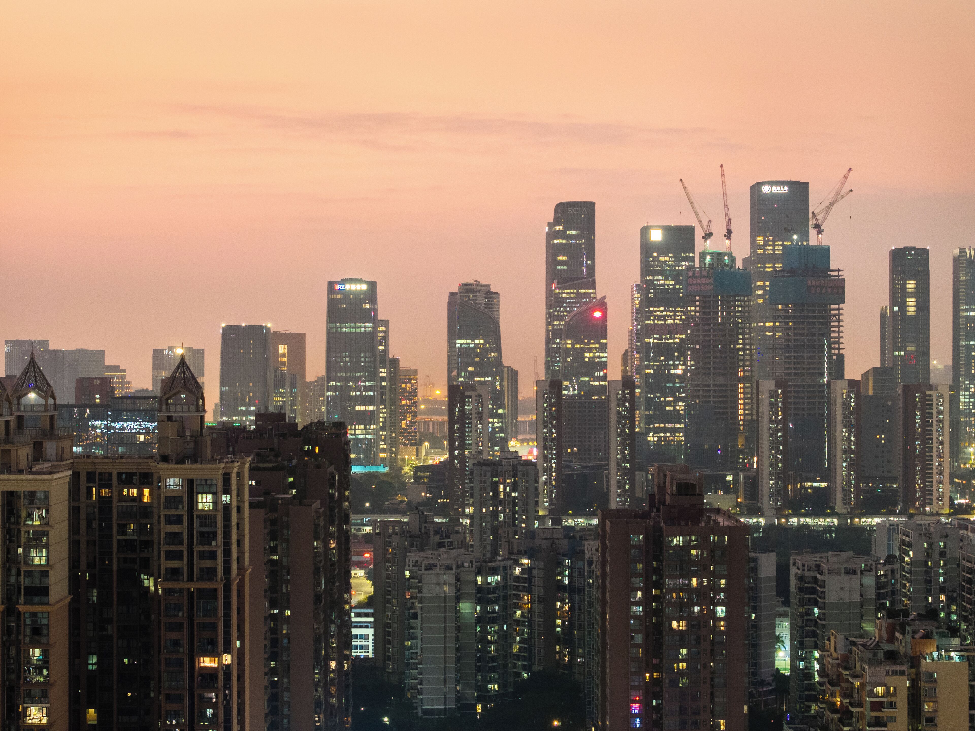 A cityscape at dusk with high-rise buildings, including a prominent SCIA building, and a construction crane visible.