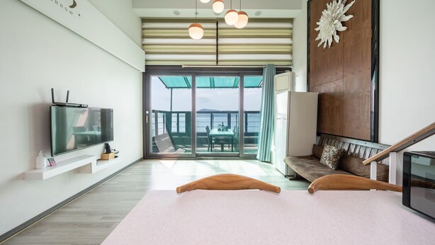 A modern living room with a flat-screen TV, a sofa, a dining table, and a view of the sea through large glass doors. - Sacheon Bichae Pension (Sacheon)