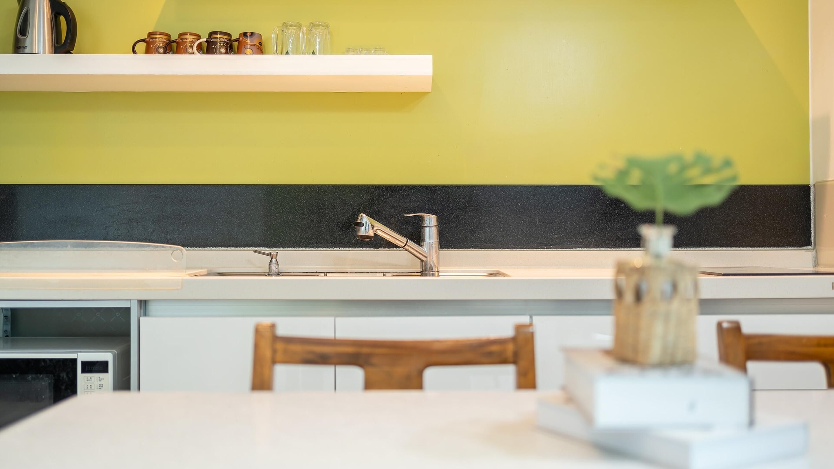 Una cocina con encimera blanca, pileta de acero inoxidable y protector contra salpicaduras negro. En primer plano, una silla de madera y una mesa blanca.