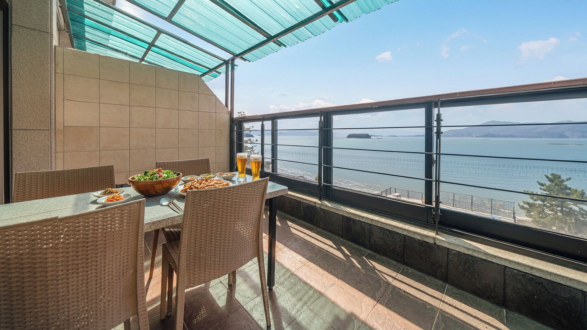 A balcony with a view of the sea, a table with food and drinks, and wicker chairs.