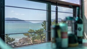 A window view of a coastal landscape with mountains in the distance, a rocky shoreline, and some greenery. - Sacheon Bichae Pension (Sacheon)