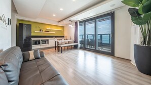 A modern living room with a sofa, a dining area with a table and chairs, a kitchen with a microwave and a stove, and a large sliding glass door leading to a balcony with a view of the sea. - Sacheon Bichae Pension (Sacheon)