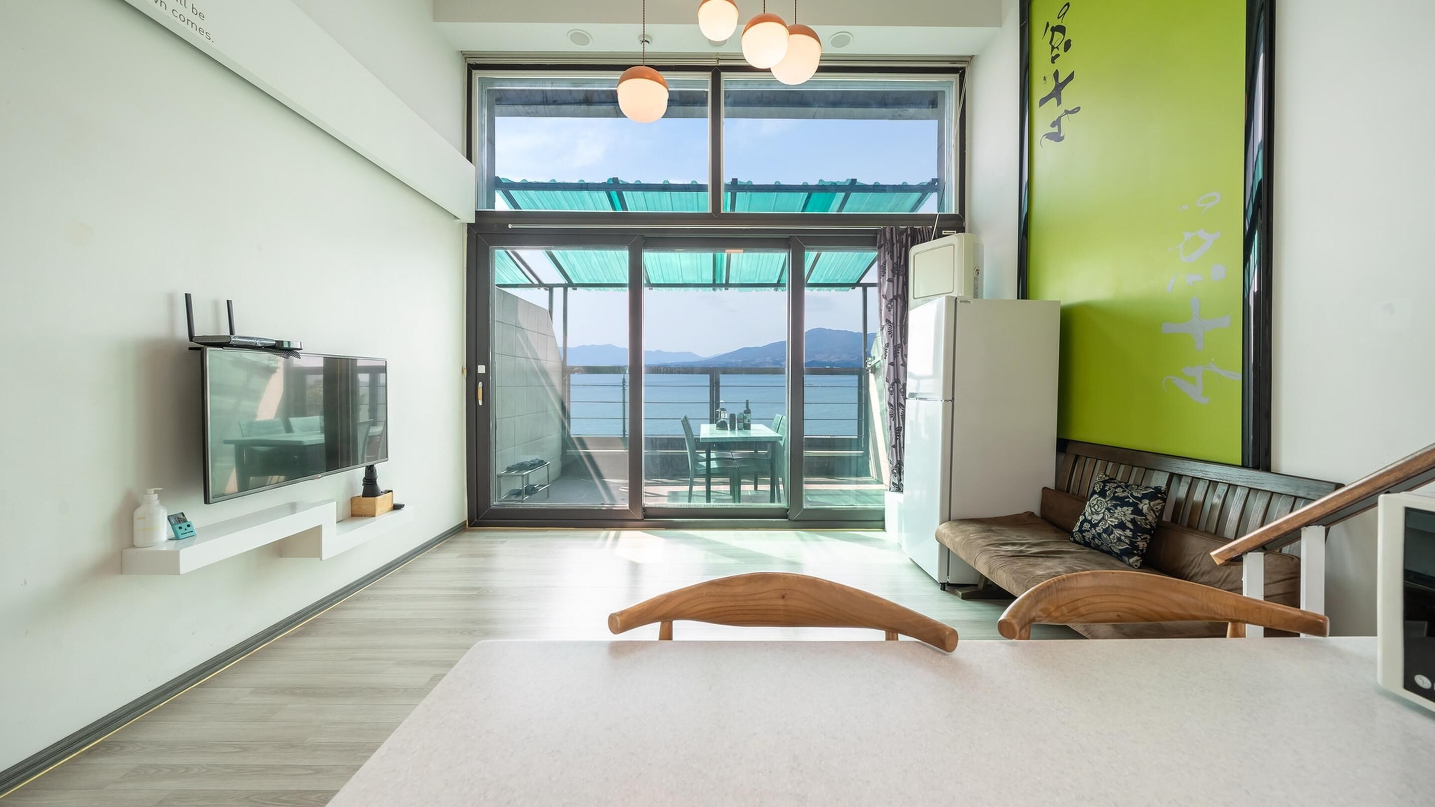A modern room with a large sliding glass door leading to a balcony with a view of the sea and mountains. There is a television on the left, a bench with a cushion, and a wooden table in the center.