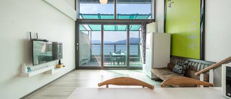 A modern room with a large sliding glass door leading to a balcony with a view of the sea and mountains. There is a television on the left, a bench with a cushion, and a wooden table in the center.