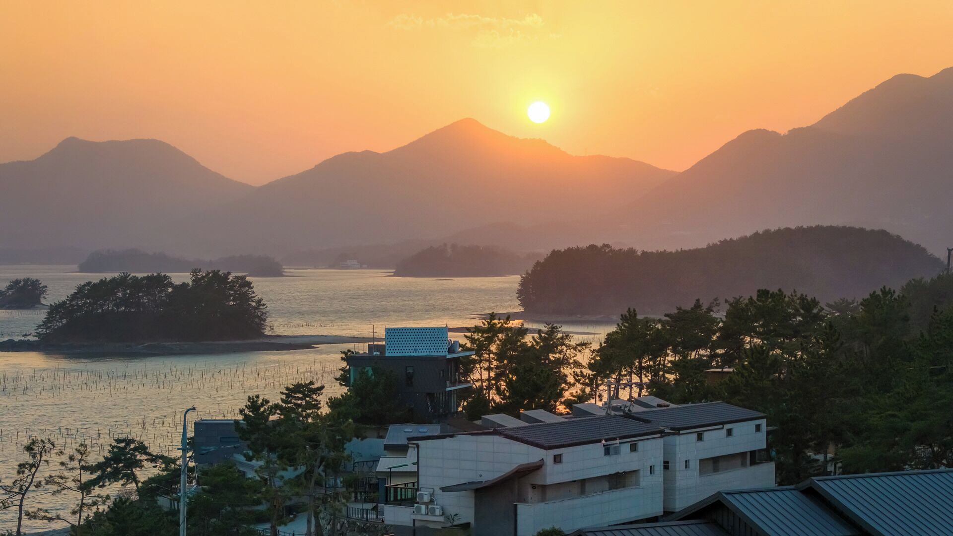 A coastal landscape with a sunset, mountains in the background, and residential buildings in the foreground.