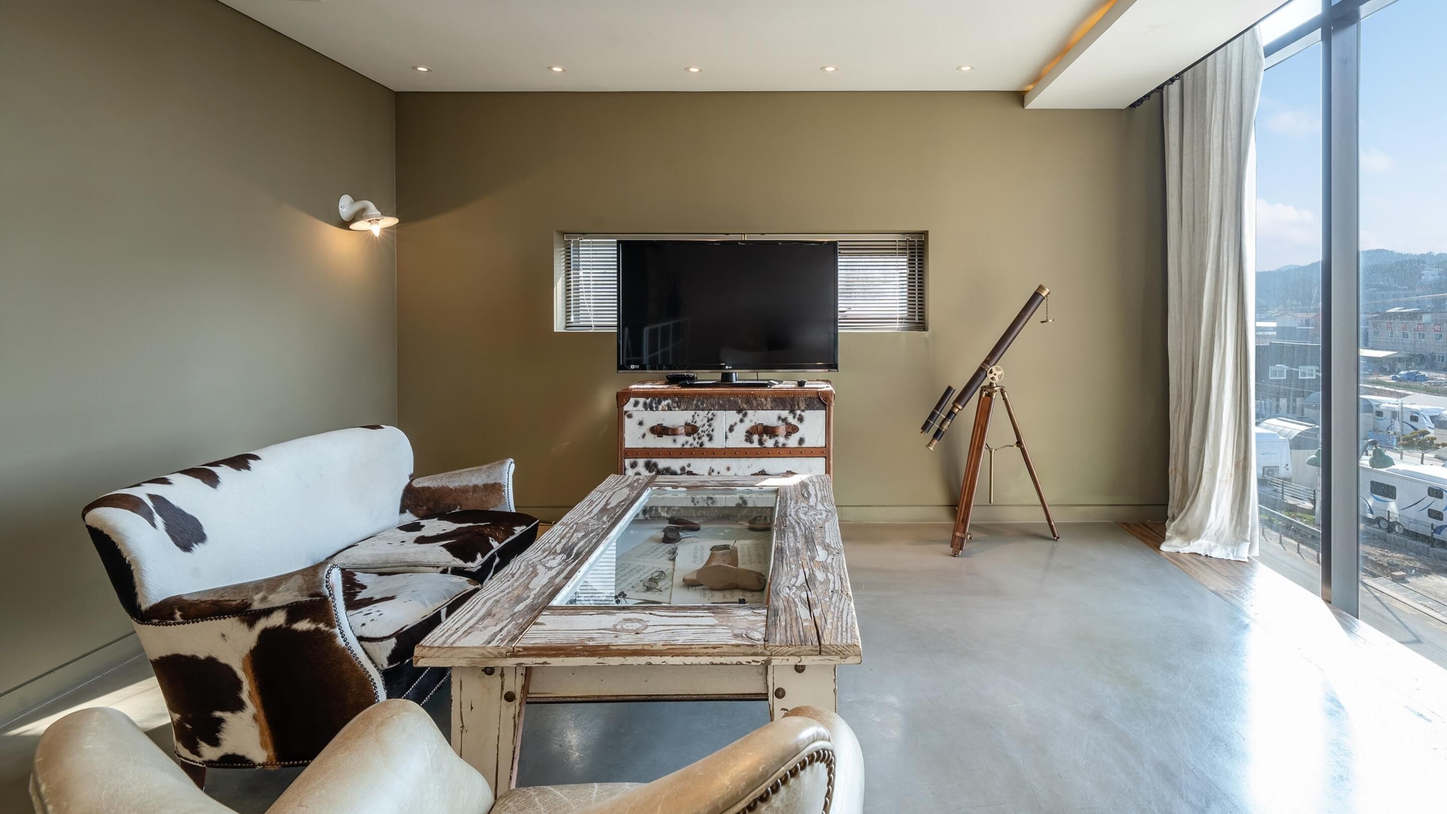 A modern living room with a large window, a mounted television, a telescope, and a unique wooden coffee table.