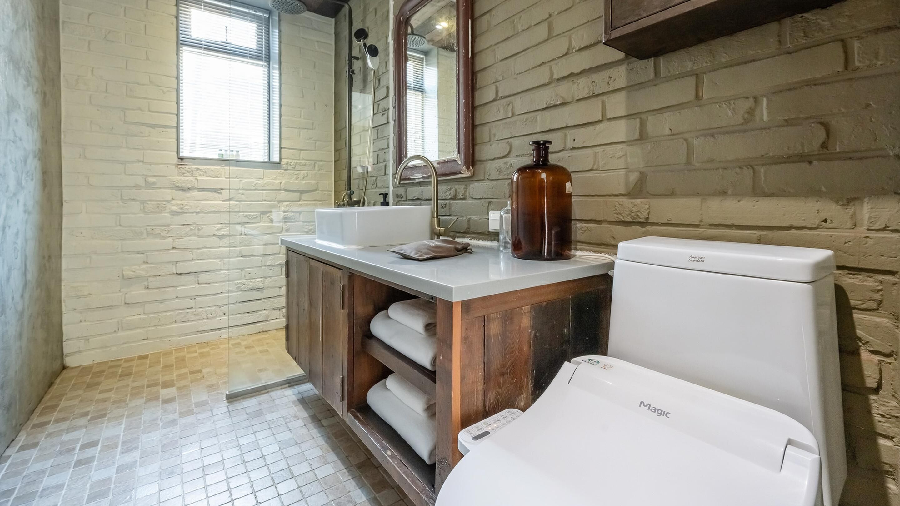 A bathroom with a white toilet, a sink, and a shower. There is a mirror above the sink and a window with blinds.