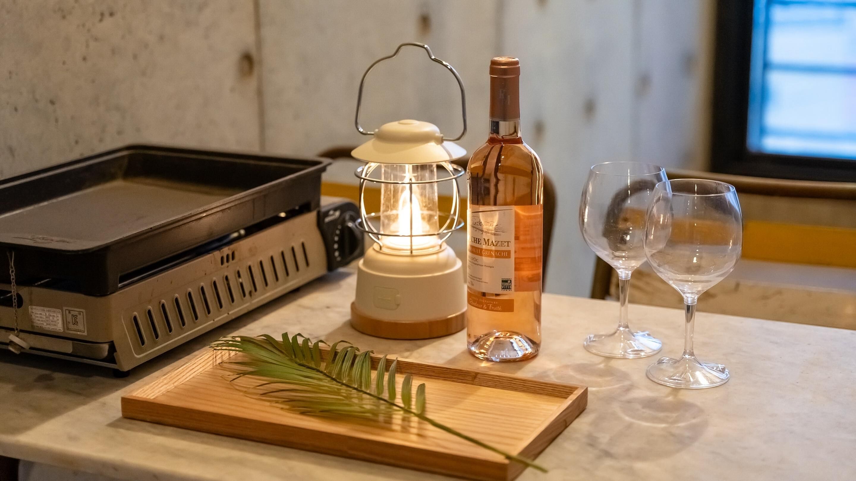 Una encimera de cocina con una hornilla portátil, un farol encendido, una botella de vino, dos copas de vino vacías y una bandeja de madera con una planta verde.