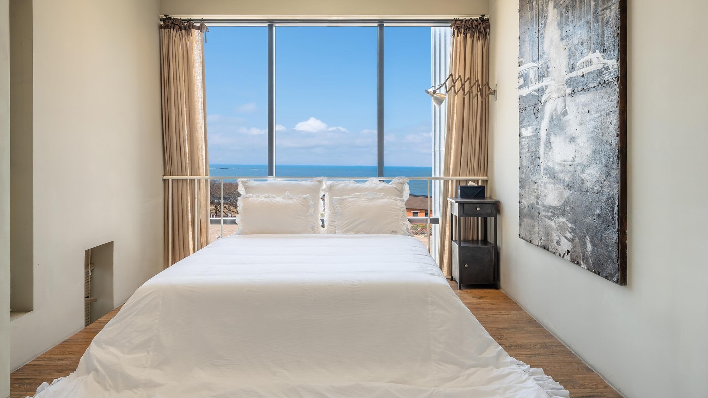 A bedroom with a large window, a bed with white linens, a wooden floor, and a modern abstract painting on the wall.