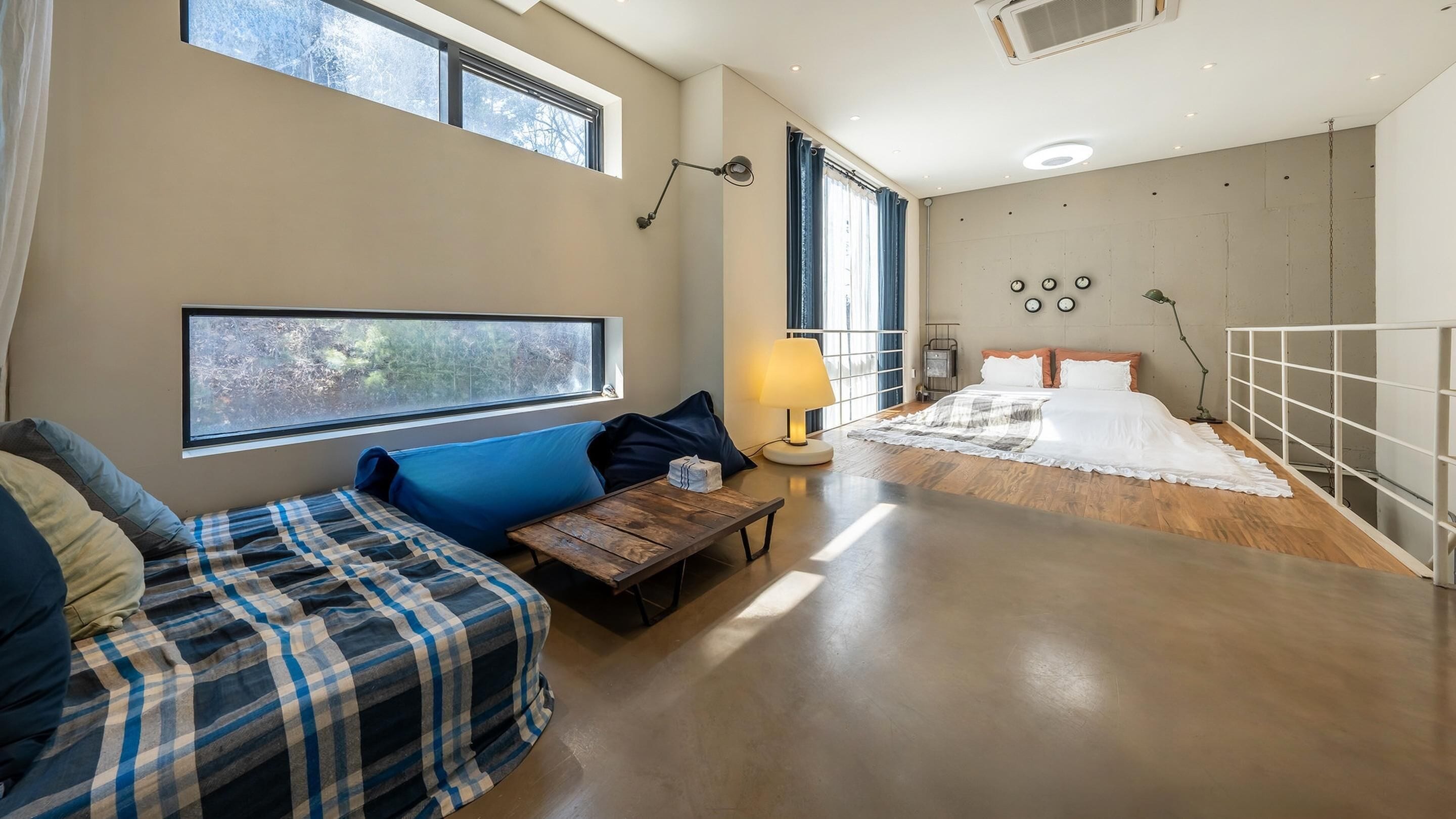 A modern bedroom with a large bed, a sofa, a wooden table, and a view of trees through the window.