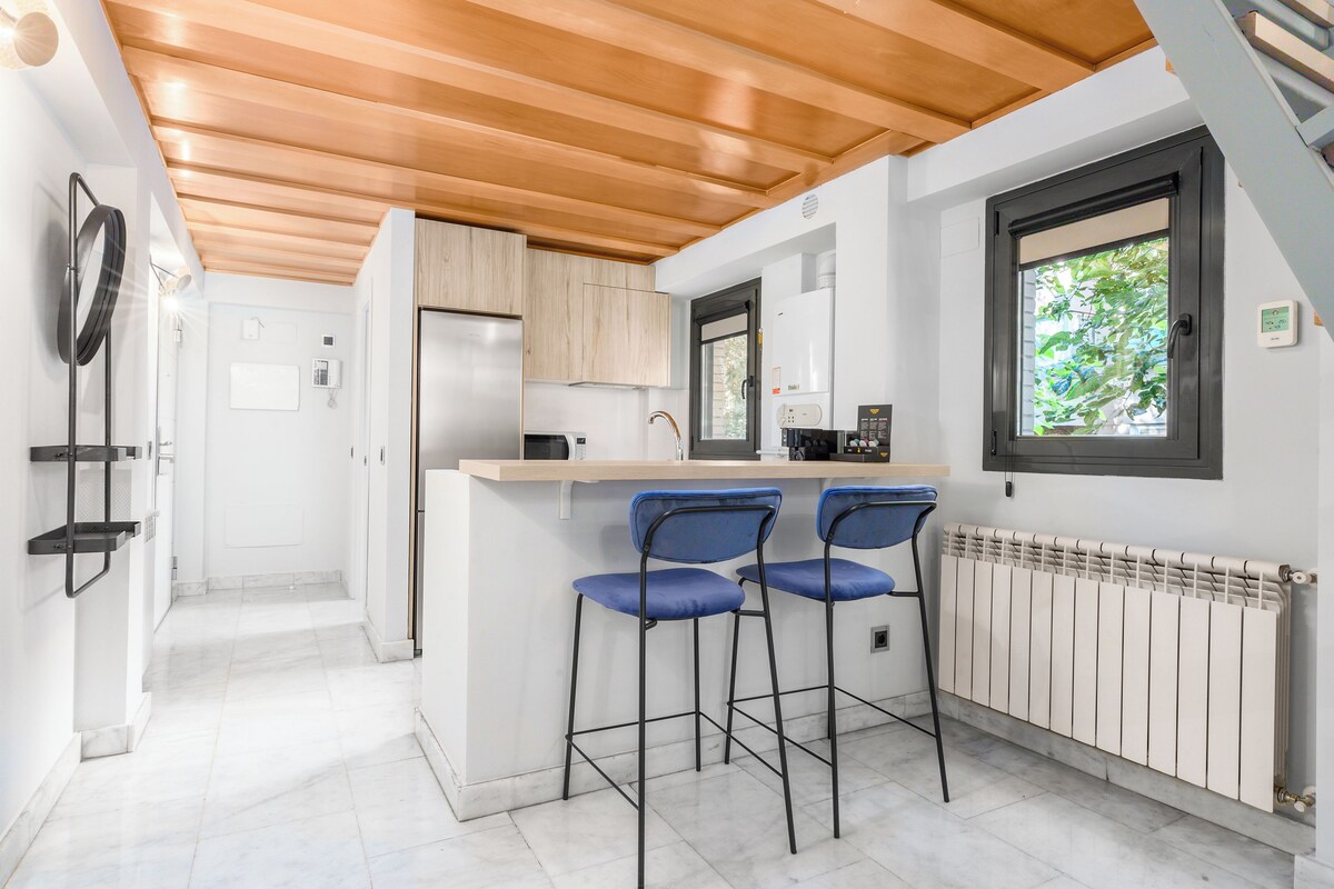Une cuisine moderne avec des poutres en bois au plafond, un plan de travail blanc, deux tabourets de bar bleus et une fenêtre donnant sur un paysage verdoyant.