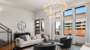 A modern living room with a sofa, armchairs, a coffee table, and a chandelier. There is a large window with a view of buildings outside. - Luxe Getaway with Hot Tub (Nashville)