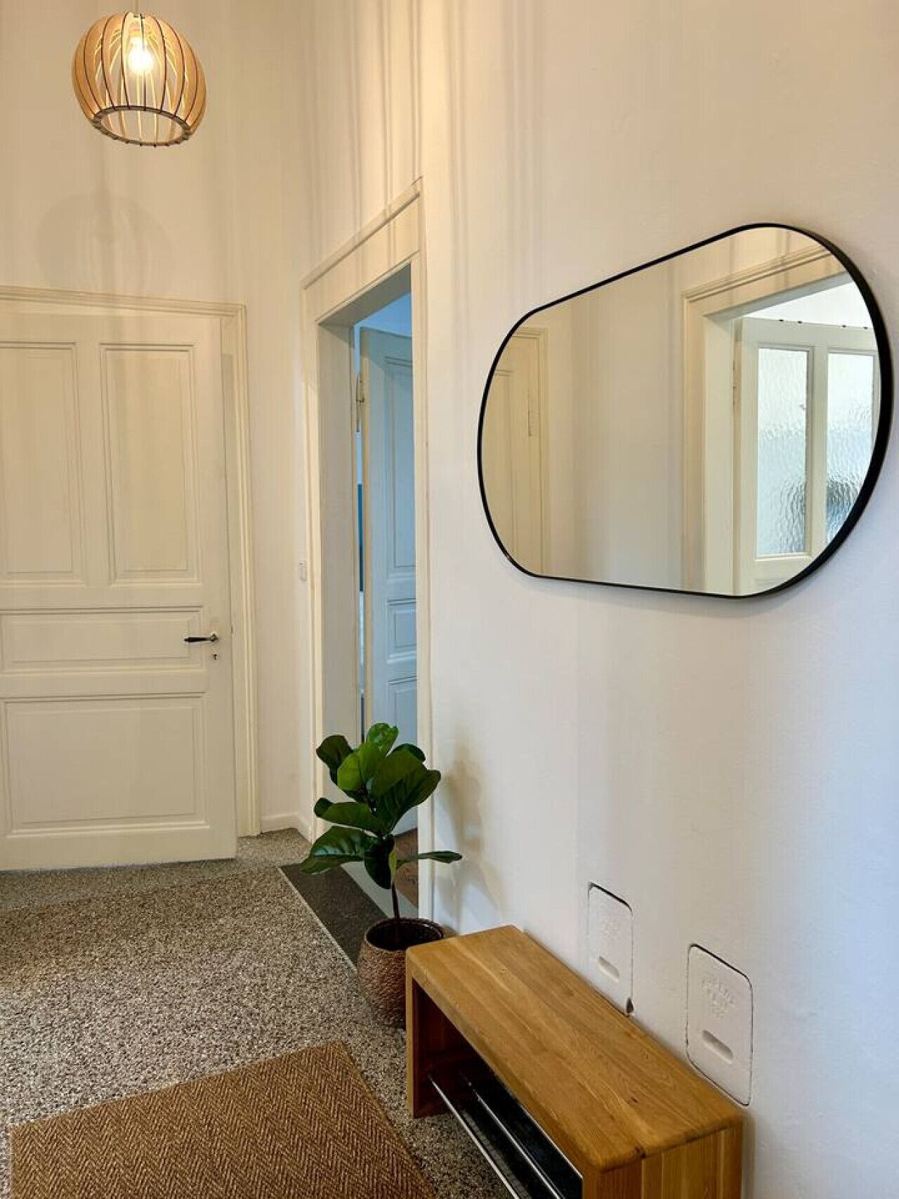 Een gang met een witte deur, een ronde spiegel, een houten bankje en een plant.