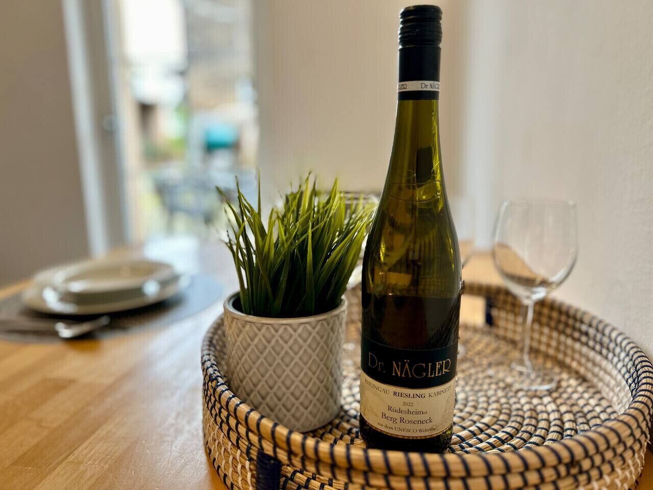 A bottle of wine, a glass, and a woven tray on a wooden table with a potted plant.