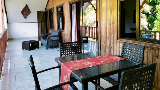A covered outdoor area with a wooden wall, a glass table with a red tablecloth, and wicker furniture. - La Source Self Catering (La Digue)