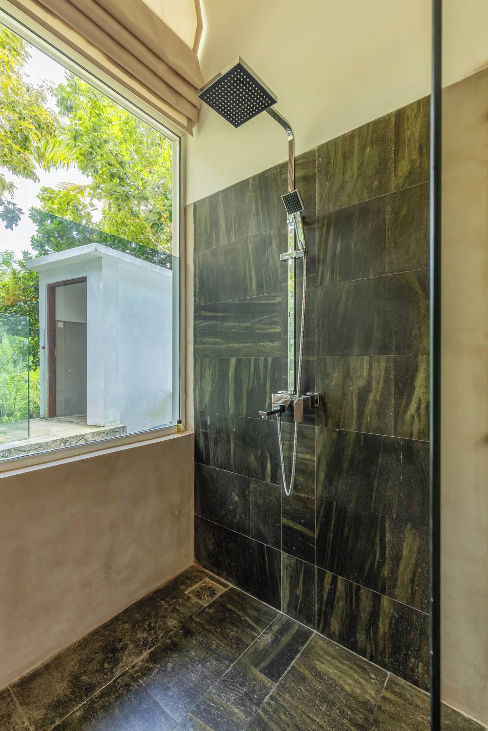 A modern bathroom with a glass shower enclosure, dark marble tiles, and a window overlooking greenery.