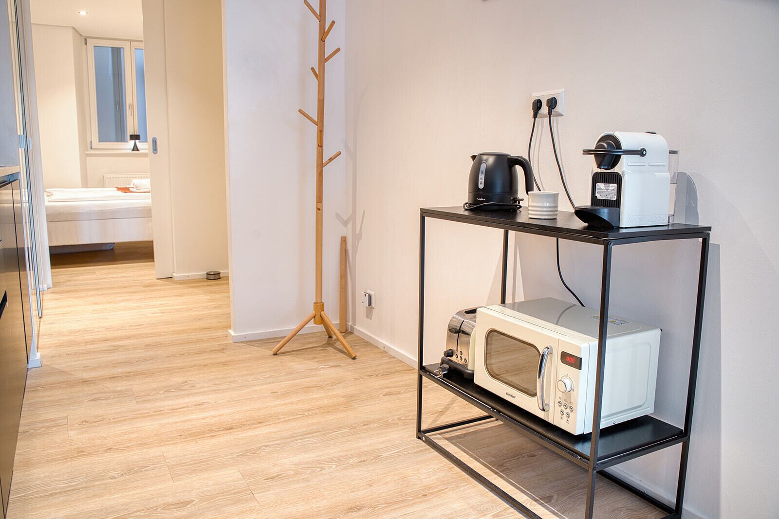 A modern kitchen with a microwave, coffee maker, and toaster on a black metal shelf. A wooden coat rack is visible in the background.