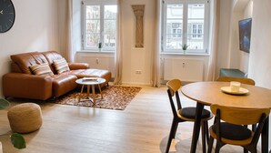 A modern living room with a leather sofa, a round wooden dining table, and two windows with curtains.