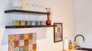 A kitchen with a shelf holding cups, a vase with a plant, a colorful tile backsplash, a poster, and a sink with a faucet.