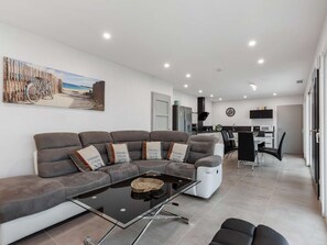 A modern living room with a sectional sofa, a glass coffee table, and a dining area with a kitchen in the background.