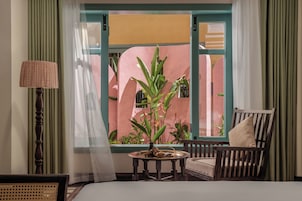 A room with a bed, a wooden chair, a table with a plant, and a window with curtains.