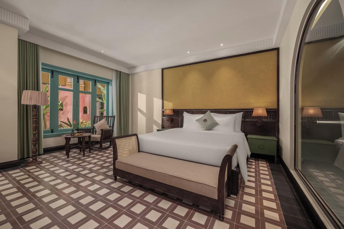 Ein Hotelzimmer mit einem großen Bett, einem Schlafsofa, einem kleinen Tisch und einem Sessel. Ein Fenster mit Vorhängen bietet Blick ins Grüne.