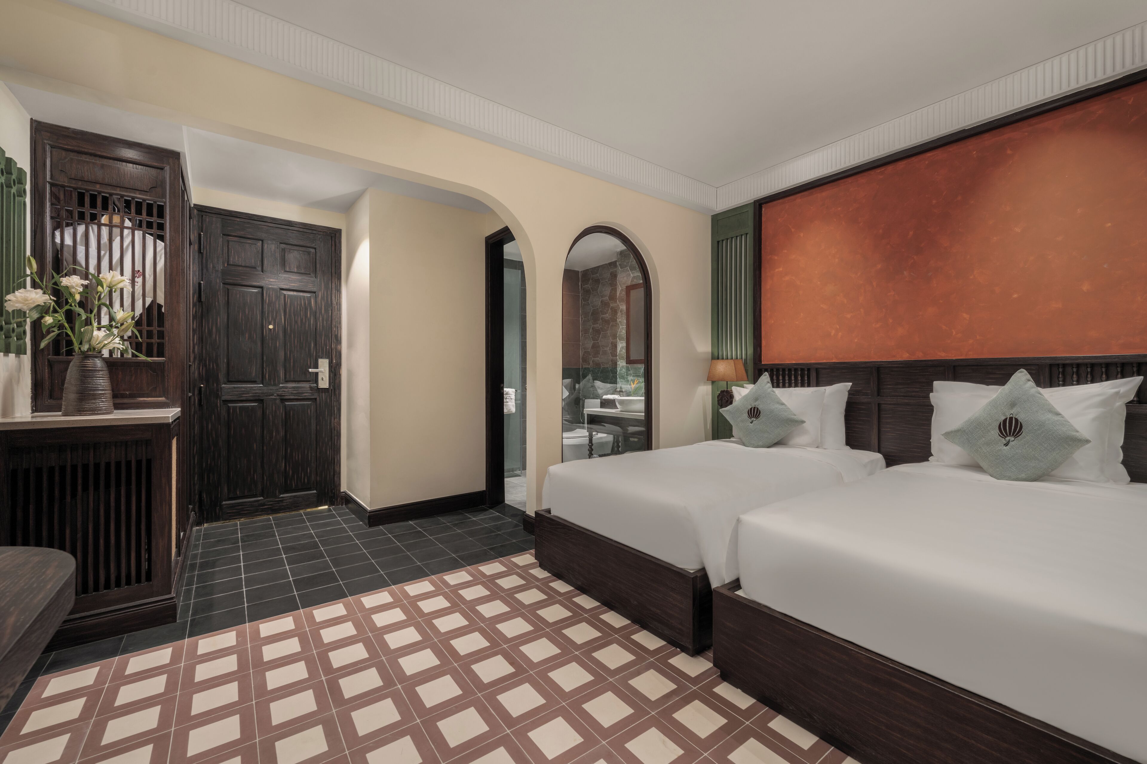 Une chambre d’hôtel avec deux lits, une armoire en bois, un sol carrelé et une vue sur l’extérieur, visible à travers une porte en arc de cercle.