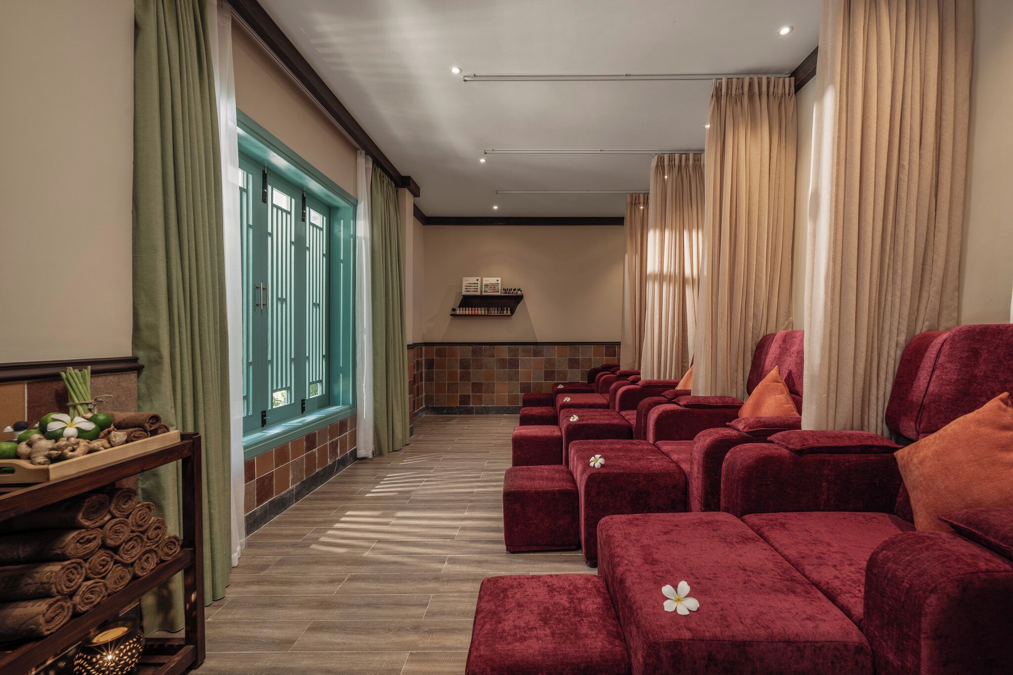 A spa room with red cushioned seating, a wooden shelf with towels and a flower, and a window with curtains.