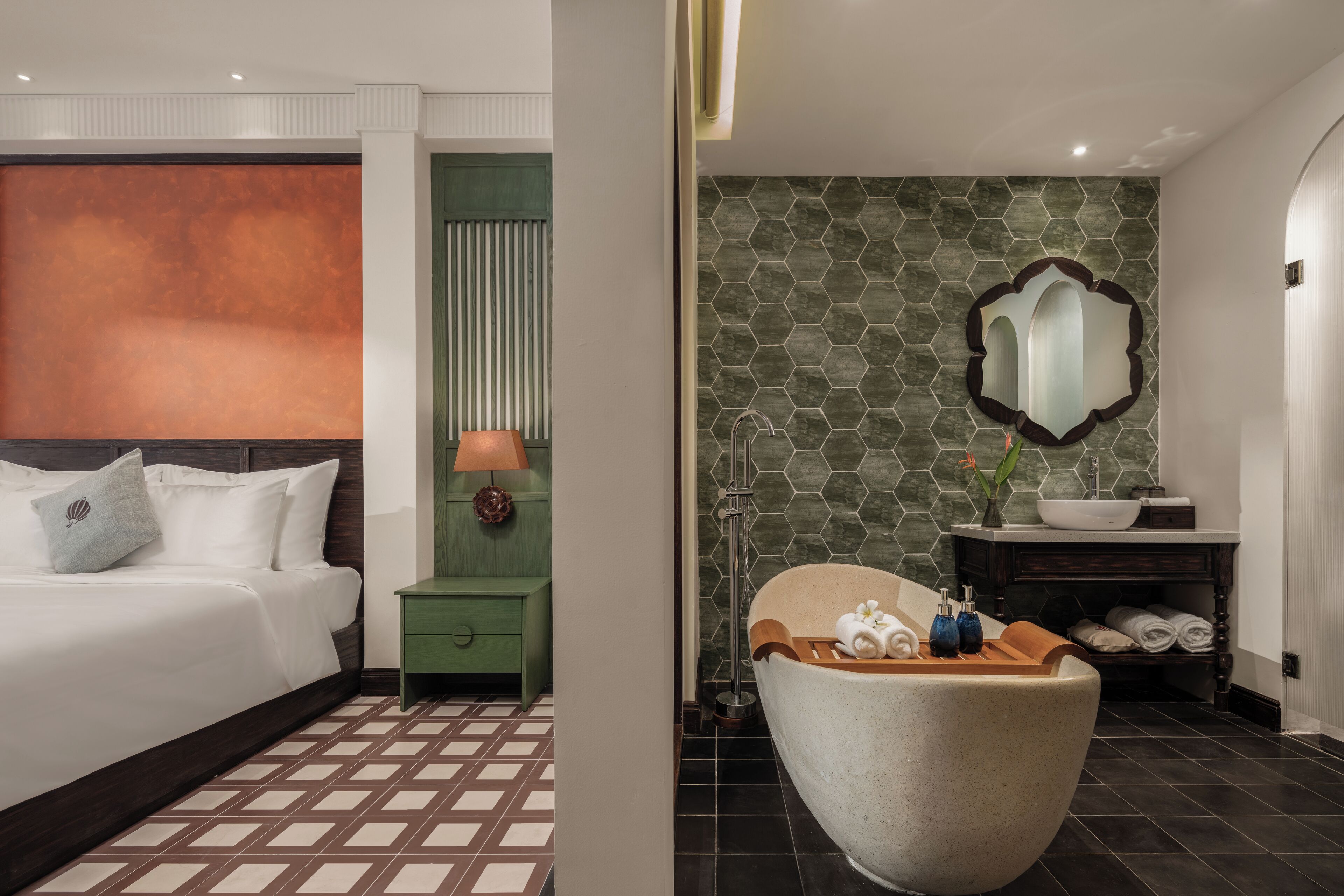 A modern hotel room with a large bed, a green nightstand, a bathroom with a freestanding bathtub, and a hexagonal patterned wall.