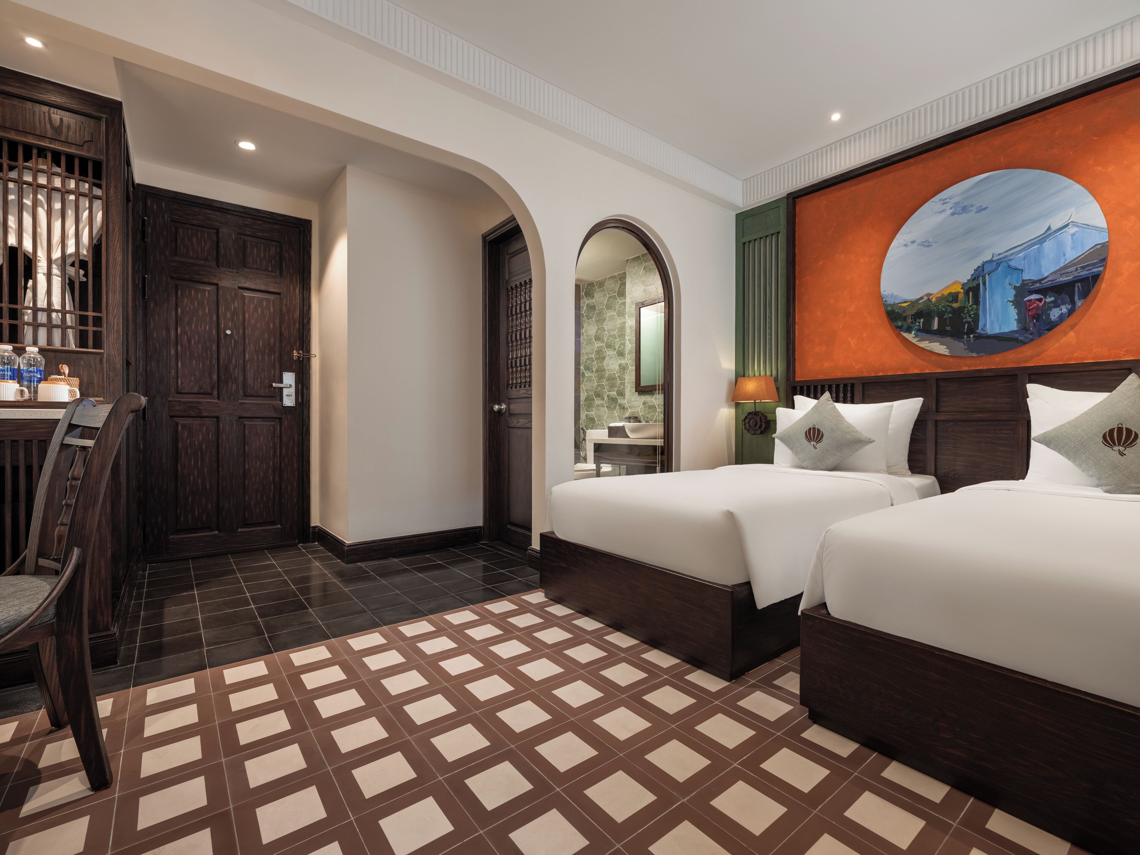 A hotel room with two beds, a desk, and a chair. There is a large painting on the wall and a tiled floor.