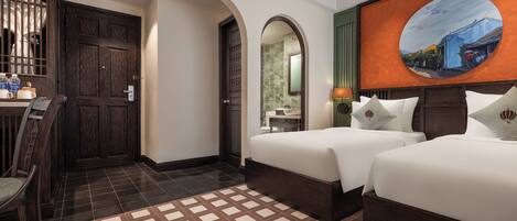 A hotel room with two beds, a desk, and a chair. There is a large painting on the wall and a tiled floor.