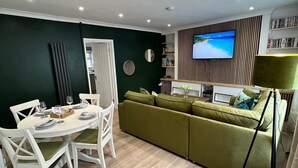 A modern living room with a green sofa, a dining table with chairs, a flat-screen TV mounted on the wall, and a lamp.