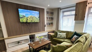 A modern living room with a green sofa, a wooden coffee table, a flat-screen TV mounted on a wooden panel wall, and a bookshelf with books and decorative items.