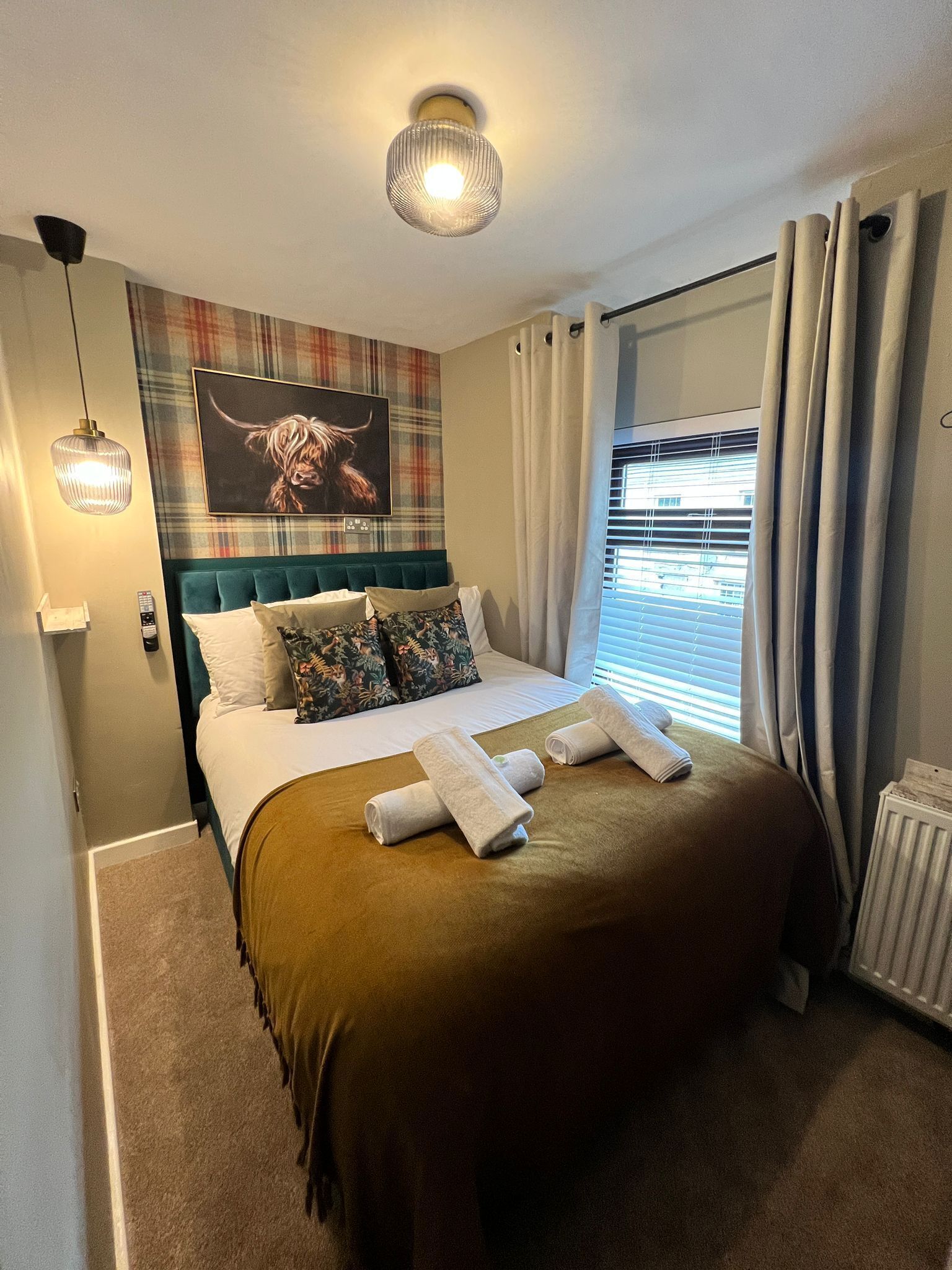 A neatly made bed with a brown bedspread and two rolled towels, a plaid-patterned wall hanging above the bed, a window with curtains, and a radiator on the floor.