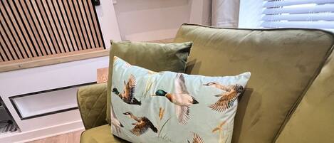 Un canapé vert agrémenté d’un coussin orné d’oiseaux et de feuilles, une bibliothèque contenant des DVD et un mur recouvert de lambris en bois.