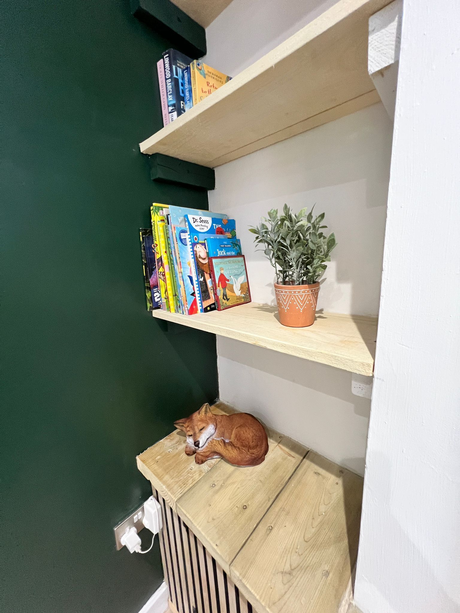 A corner of a room with a green wall, a wooden shelf with books and a potted plant, and a small wooden table with a sleeping dog.