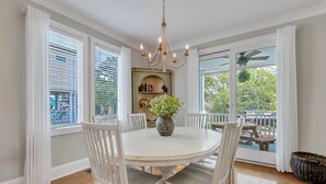 Un espace repas avec une table ronde, des chaises blanches, un lustre et une vue sur l’extérieur grâce à de grandes fenêtres.