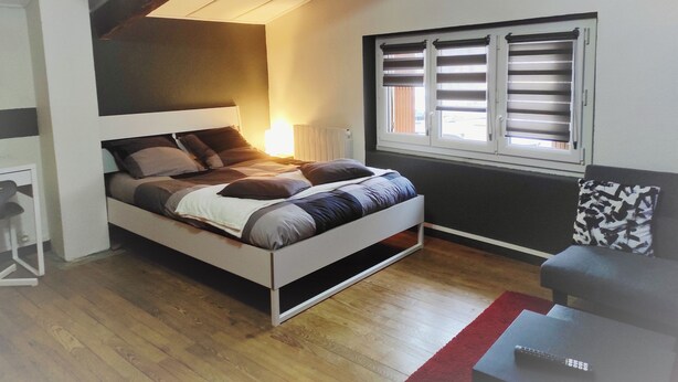 A bedroom with a bed, a window with blinds, a radiator, and a small table with a remote control. - Studio Duplex Avec Terrasse Ombragée (Lempdes)
