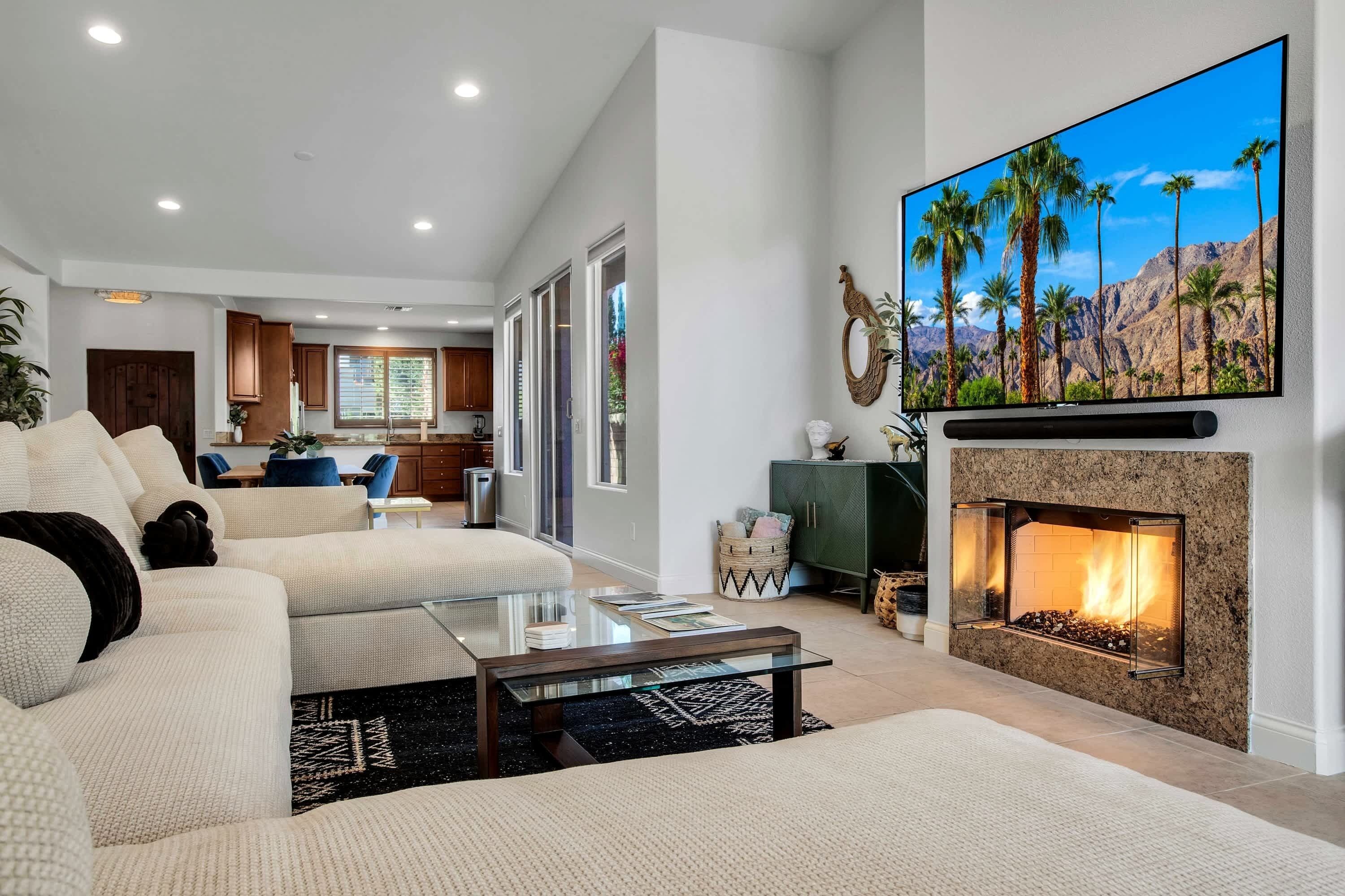 A modern living room with a fireplace, a large flat-screen TV, and a kitchen in the background.