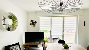 Ein modernes Wohnzimmer mit Fernseher, Esstisch, Stühlen, einer Pendelleuchte und einem großen Fenster mit Jalousien.