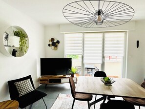 Ein modernes Wohnzimmer mit Fernseher, Esstisch, Stühlen, einer Pendelleuchte und einem großen Fenster mit Jalousien.