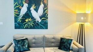 Ein beiges Sofa mit zwei Zierkissen, ein an der Wand hängendes Bild von weißen Vögeln und eine Stehlampe mit gemustertem Lampenschirm.