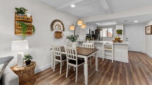Un coin repas avec une table en bois et des chaises blanches, une petite table d’appoint en osier avec une lampe, et une étagère murale pour les plantes.