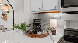 Une cuisine avec des meubles blancs, une machine à café noire, un plateau en bois avec des bocaux et une lampe suspendue.
